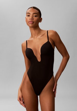 Femme portant un maillot de bain une pièce noir avec un décolleté en V profond et de fines bretelles, debout devant un fond gris uni.