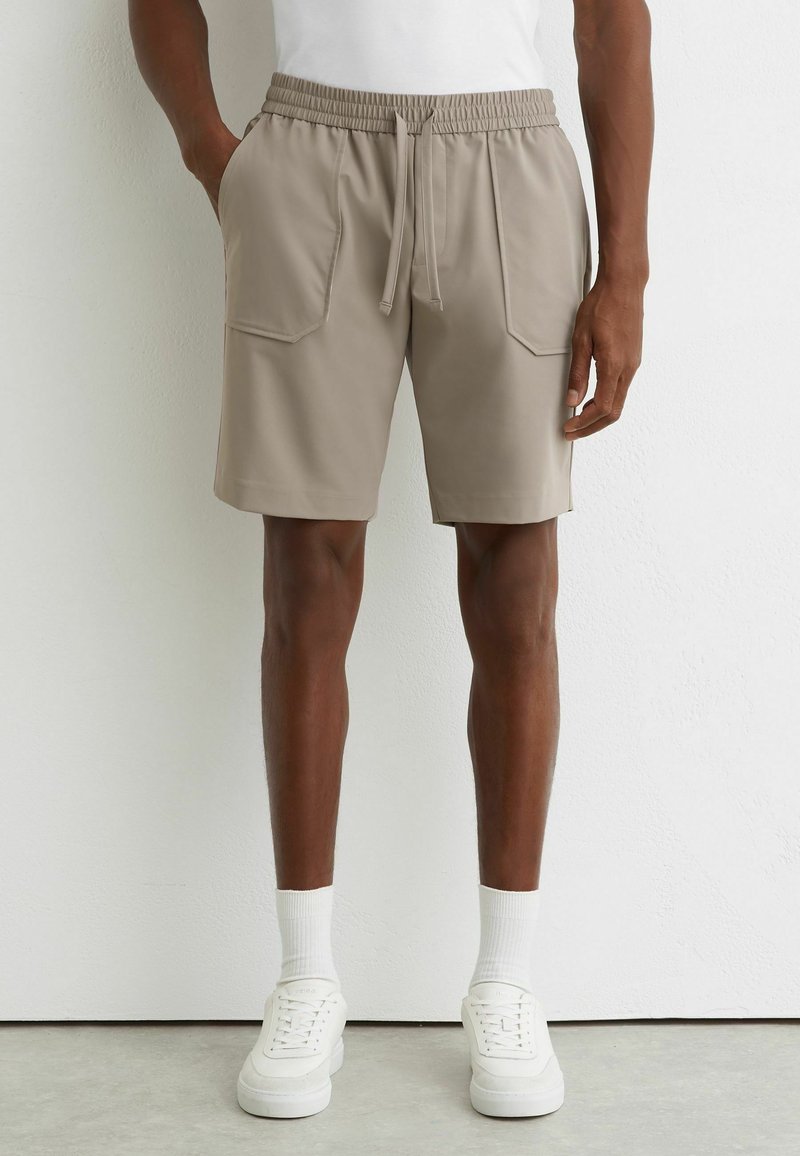 Homme portant un short beige mi-cuisses avec des poches avant, des chaussettes blanches, des baskets blanches et une chemise blanche, debout contre un mur uni.