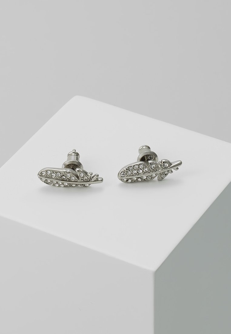 Boucles d’oreilles clous couleur argent en forme de plumes, ornées de petits strass clairs, posées sur une surface blanche.