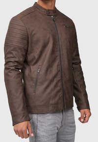 Veste en faux cuir marron au design ajusté, avec des accents côtelés sur les épaules, des poches zippées et un col haut. Détails de couture visibles.