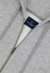 Jasnoszara bluza z kapturem na zamek błyskawiczny o teksturowym wykończeniu, z frontowym zamkiem, białą podszewką oraz metką z logo blisko kołnierza.