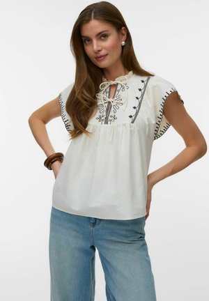 Młoda kobieta z długimi brązowymi włosami nosi białą haftowaną bluzkę i jasnoniebieskie dżinsy, pozując z rękami na biodrach na białym tle.