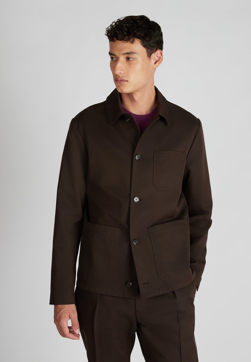 Brązowa robocza kurtka z kołnierzem, frontowymi guzikami i dwoma kieszeniami naszytymi. Tkanina ma fakturę, a w designie zawarte są subtelne detale.