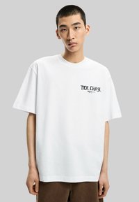T-shirt bianco in cotone con maniche corte, caratterizzato da un logo stampato nero "TIDE CHASE" nella zona sinistra del petto. Design semplice e oversize.