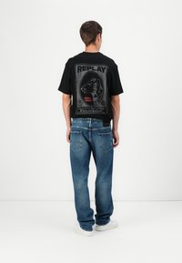 Black t-shirt with a graphic print on the back featuring an animal design and "REPLAY" text. Loose-fitting blue denim jeans and white sneakers.