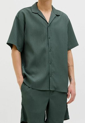 Homme portant une chemise à manches courtes côtelée vert foncé boutonnée et un short côtelé assorti, debout les mains dans les poches.