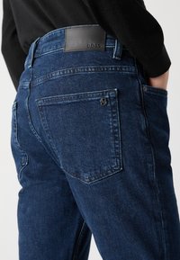 Temno modri kavbojki iz denima, ki imajo usnjeno nalepko z blagovno znamko "BOSS", zadnje žepke in gladko, rahlo teksturirano tkanino.