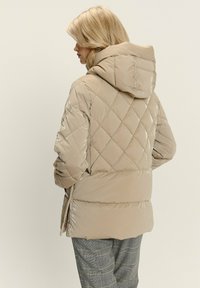 Giacca a vento imbottita beige con cappuccio, caratterizzata da una superficie lucida, cerniere laterali e motivi a rombo sul tessuto.