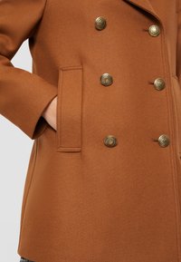 Bruine dubbelgeknoopte jas met textuurstof, gouden knopen en zijzak. Perfect gesneden met gekartelde revers.