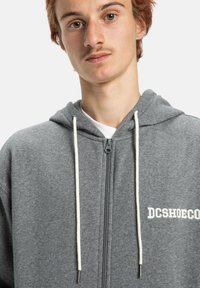 Grijze zip-up hoodie van zacht materiaal, met een trekkoordcapuchon, witte accenten en het geborduurde "DCSHOECO" logo op de voorkant.