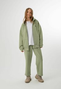 Ljusgrön zip-up hoodie med stora fickor, tillsammans med matchande sweatpants. Vit t-shirt under; modellen bär beige sneakers.