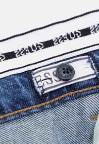 Ceinture de pantalon en denim avec un bouton noir, une bande blanche portant le texte 'Guess' et une étiquette indiquant la taille en bleu et blanc.