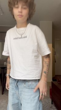 Camiseta blanca corta con el texto "abejas y chucherías", combinada con pantalones cortos de mezclilla anchos de color azul claro. Se pueden ver pulseras de plata y tatuajes.