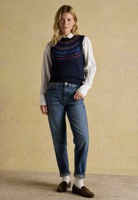 Jūras spēku adīts vests ar daudzkrāsainām ģeometriskām rakstām, valkāts virs balta krekla ar apkakli, kombinēts ar ziliem džinsiem un brūniem loafers.