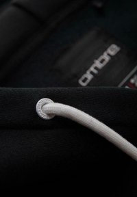 Nahaufnahme eines weißen Kapuzenpullischnürzugs, der durch ein metallisches Ösenloch auf schwarzem Stoff gefädelt ist, mit einem verschwommenen "ambro"-Label im Hintergrund.