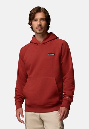 Homme aux cheveux châtain clair portant un sweat à capuche rouge Columbia et un pantalon beige, debout devant un fond clair uni.