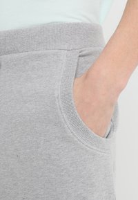 Ręka umieszczona w przedniej kieszeni jasnoszarych spodni dresowych z ribbowanym paskiem w talii i wykończeniem kieszeni.