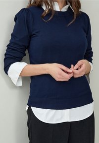 Granatowy sweter dzianinowy z bufkami, z prążkowanymi mankietami i dolną krawędzią, noszony na białej koszuli z kołnierzykiem. Teksturowany materiał i dopasowany krój.