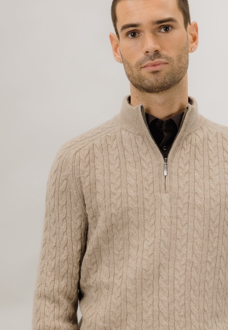Beige kabelstrikket genser med høy krage og kvart glidelås. Myk tekstur; subtile detaljer i strikkemønsteret.