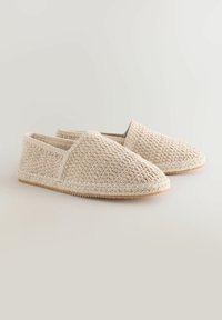 Zapatos slip-on de tejido beige con superficie texturizada, puntera redondeada y detalle de cuerda de yute a lo largo de la suela. Diseño ligero y casual.