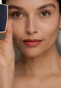 En kvinne som holder et rektangulært, mørkeblått kompakt speil med en gullkant, som viser glatt hud og en myk, naturlig leppestiftfarge.