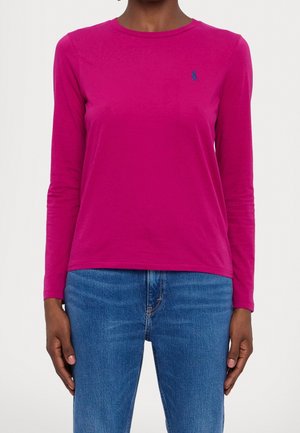Vrouw draagt een felroze shirt met lange mouwen met een klein blauw geborduurd logo op de linkerborst en een blauwe spijkerbroek.