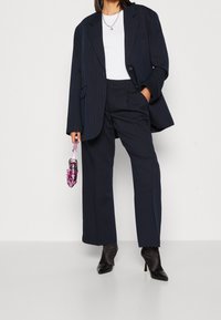 Abito a righe navy caratterizzato da un blazer oversize, pantaloni dritti e un top bianco, abbinato a tacchi a punta neri e un ombrello rosa.