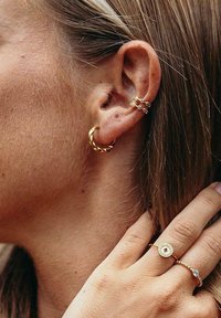 Goldene Creolen mit verdrehtem Design, kombiniert mit einem gestapelten Ohrcuff, der kleine Steine enthält. Ein goldener Ring mit kreisförmigem Akzent an einer Hand.