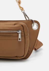 Beige stoftas med en sølvfarvet lynlås-lomme, rund øje, og et snoet plastik håndtag. Blød tekstur med et elegant, vinklet design.