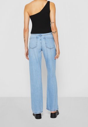 Woman with tattoos wearing a black one-shoulder top, light blue high-waisted jeans, and black platform shoes, standing facing away.