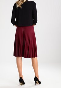 Black three-quarter sleeve top paired with a knee-length, pleated burgundy skirt and black stilettos, shown from the back.