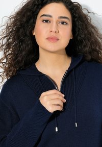 Mörkblå huvtröja med dragkedja i halsen, med ribbad detaljer och snoddar med metallspetsar. Mjuk textur, avslappnad passform.