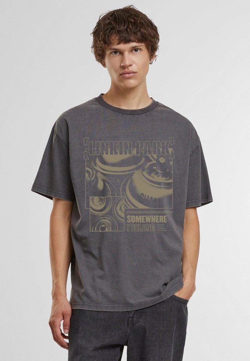 Jeune homme aux cheveux bouclés portant un t-shirt gris foncé délavé Linkin Park avec le texte "Somewhere I Belong" et des graphismes abstraits.