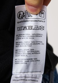 Verzorgingslabel op stof met meerdere talen. Text geeft aan: 100% katoen, wasinstructies en zorgsymbolen op een witte achtergrond.