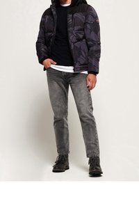 Vadderad jacka i mörkt kamouflage-mönster med en svart axelpanel, kombinerad med grå jeans och svarta stövlar. Har en huva.