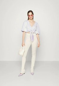 Blusa curta de cor lilás claro com mangas bufantes e detalhe de botão, combinada com calças brancas de cintura alta e sapatos lilás com bico fino, segurando uma pequena bolsa.