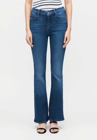 Γυναίκα που φοράει μπλε τζιν παντελόνι μεσαίου ύψους και καμπύλο τελείωμα στα μπατζάκια και μαύρα σανδάλια, στέκεται μπροστά σε ένα απλό φωτεινό φόντο.