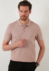 Világos rózsaszín pólóing gallérral, három gombos nyitással, rövid ujjal és texturált anyaggal. Fekete nadrággal és órával viselve.