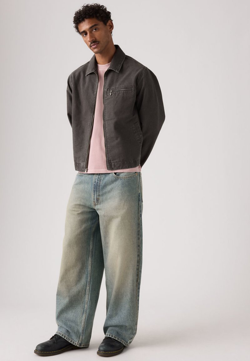 Mørkegrå lynlåsjakke over en lyserød t-shirt, kombineret med vide, falmede blå jeans og sorte sko.