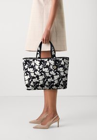 Torebka typu tote w czarno-białym wzorze kwiatowym, z dwiema solidnymi rączkami i okrągłym akcentem ze stali. Modelka ma na sobie spódnicę w kratę.