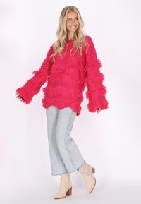 Fuchsia franjesweater met brede mouwen, gecombineerd met lichtblauwe flared jeans en beige enkellaarsjes, op een model tegen een neutrale achtergrond.
