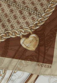 Sciarpa con sfondo beige a motivo, dettagli marroni, design a catena ornamentale e un charm a forma di cuore dorato con la scritta "GUESS".
