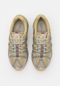 Sportskor med en beige och grå mesh-överkonstruktion, ljusblå snören och en dämpad sula med en framträdande häl.