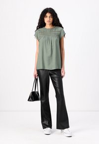 Blouse vert clair avec une taille froncée et des détails en dentelle, associée à un pantalon évasé en simili cuir noir et des baskets blanches. Sac à main noir inclus.