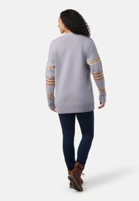 Ljuslila tröja med mönstrade orangea och gula ränder på ärmarna, texturerad stickad tyg, oversized passform, kombinerad med mörka jeans och bruna stövlar.