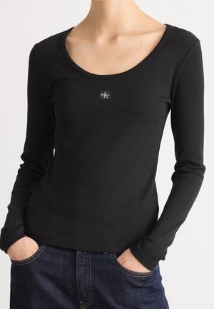 Zwart geribd topje met lange mouwen en een ronde halslijn, met een klein Calvin Klein-logo op de borst, gecombineerd met donkere denim jeans met handen in de zakken.
