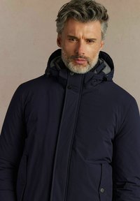 Granatowa, ocieplana kurtka z kapturem, zatrzaskami i bocznymi kieszeniami. Ma teksturowane wewnętrzne wykończenie oraz elegancki, nowoczesny design.