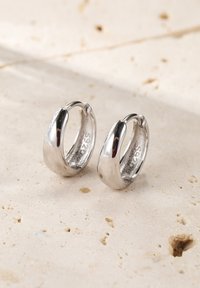 Ohrringe aus Sterlingsilber mit einer glatten, polierten Oberfläche, die eine gebogene Form und ein offenes Verschlussdesign aufweisen. Auf einer strukturierten beigen Oberfläche präsentiert.