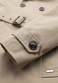 Beżowy trench coat z rękawem z guzikami i detalem w postaci patki. Wykonany z mocnego materiału o gładkiej fakturze, z metalowym akcentem w postaci klamry.