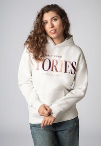 Crème hoodie met "ENKEL GOEDE VERHALEN" in grote, gewatteerde letters gedrukt. Heeft een voorvak en geribbelde manchetten. Combinatie met een blauwe jeans.